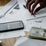 Mãos sobre uma mesa com smartphone, dinheiro, relatórios financeiros e calculadora